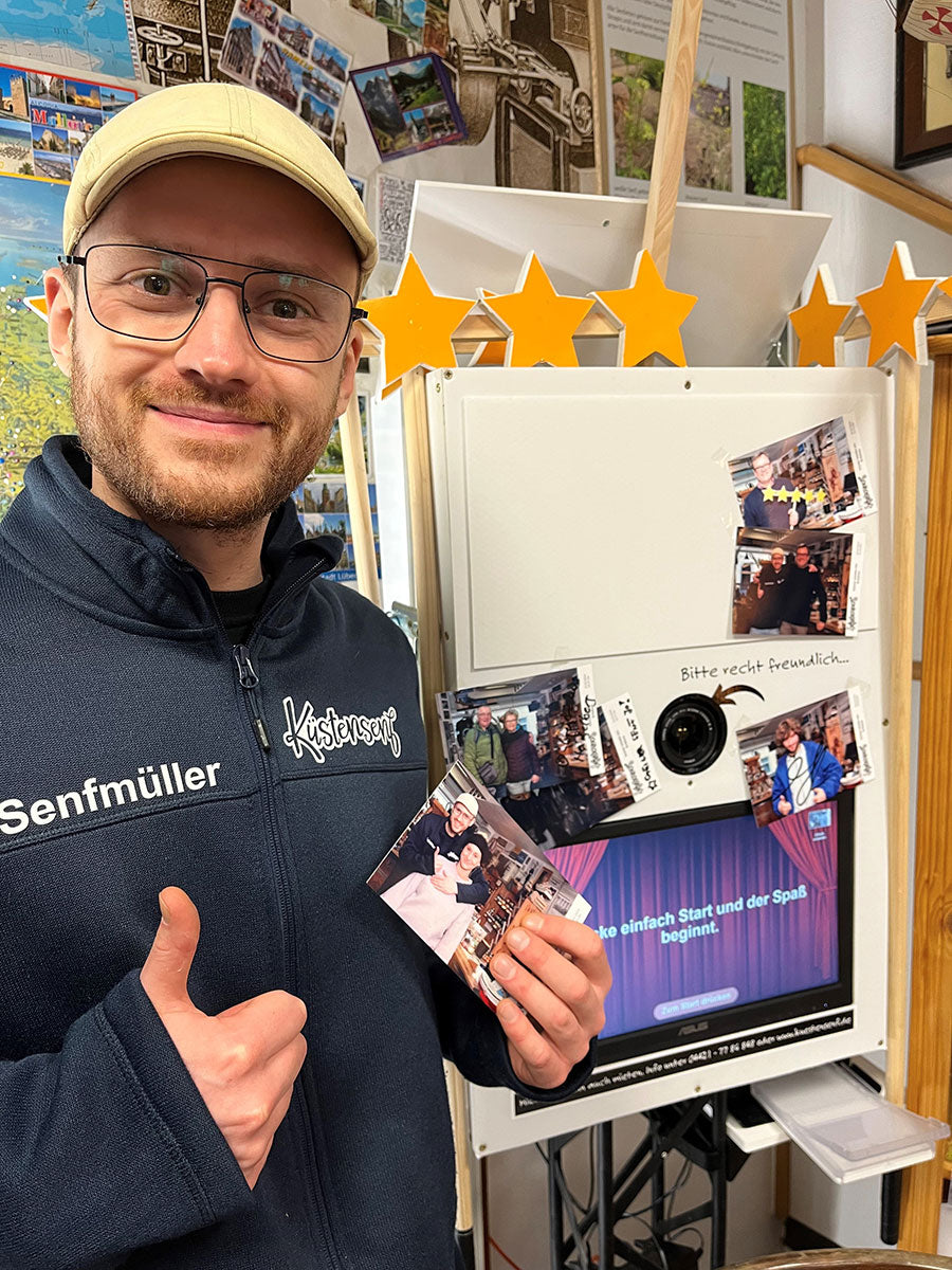 Senfmüller Andy steht vor der Fotobox. Die Fotobox ist ein weißer kasten mit einem Bildschirm und einem Monitor. Er hält ein Bild in der Hand und zeigt einen Daumen hoch.
