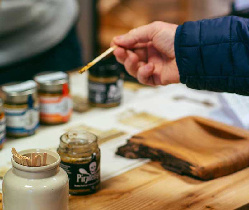 man sieht die Hand eines Kunden. Er hält einen Holzstab in der Hand, mit dem er aus einem Senfglas etwas Senf zum Probieren entnimmt.